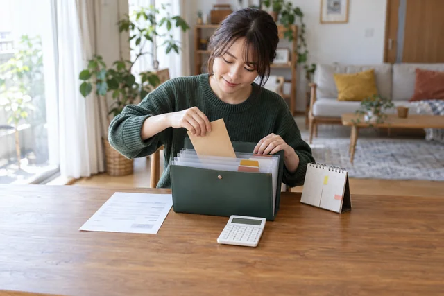 電卓と明細、付箋を貼った卓上カレンダーを前に、税金の分を封筒に分けて書類ケースへしまう女性